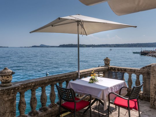 Leckeres in Eurem Hotel mit Strand am Gardasee Tisch mit Sonnenschirm und zwei Stühlen mit Blick auf einen See