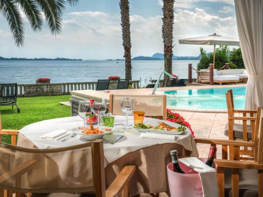 Leckeres in Eurem Hotel mit Strand am Gardasee Tisch mit Essen und Wein neben Pool und See mit Palmen