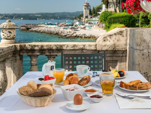 Ein charmantes Bed-and-Breakfast-Konzept in Gardone Riviera Frühstück mit Brötchen, Saft und Kaffee am Seeufer mit historischem Turm im Hintergrund