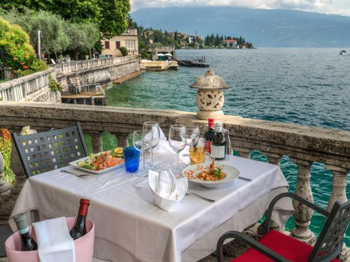Willkommen in Eurem Hotel in Gardone Riviera! Tisch mit Essen und Wein an einem Balkon mit Blick auf einen See und Berge