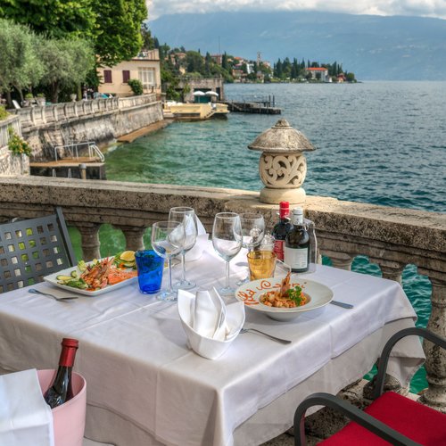 Leckeres in Eurem Hotel mit Strand am Gardasee Tisch mit Essen und Wein an einem Balkon mit Blick auf einen See und Berge